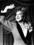 Bette Midler Portrait Waving Her Hand in Grey Linen Deep V-Neck Dress with White Ruffled Collar and Flower Corsage Photo Print (8 x 10)